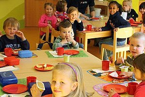 Viele Kinder an Esstischen sitzend lächeln in die Kamera – das Bild verlinkt auf die Seite 'Kindertagesstätte'