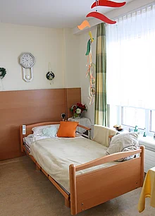 Zimmer mir einem Pflegebett, daneben große Fenster, teilweise mit Gardinen verdeckt